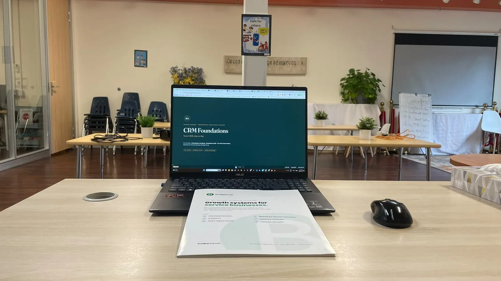 A laptop open on the first slide of a Brydge Academy course, beside the printed course workbook